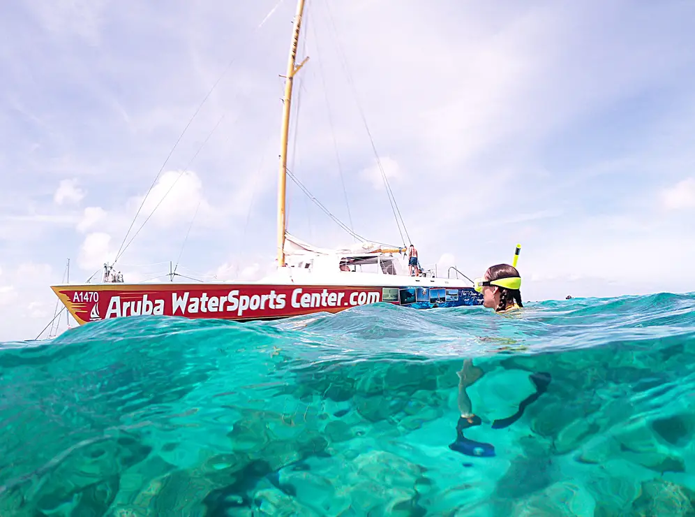 woman wearing yellow snorkel and goggles in turquoise-colored water beside Aruba Watersports Center catamaran