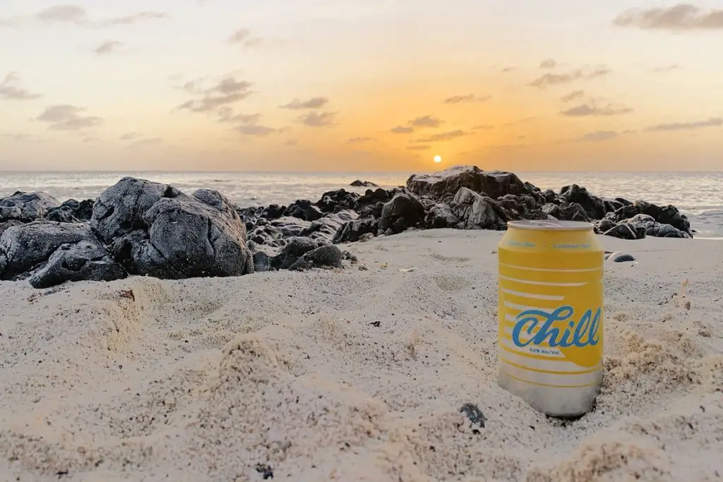 a can of Balashi Chill beer sitting on the sand in front of grey rocks at sunset