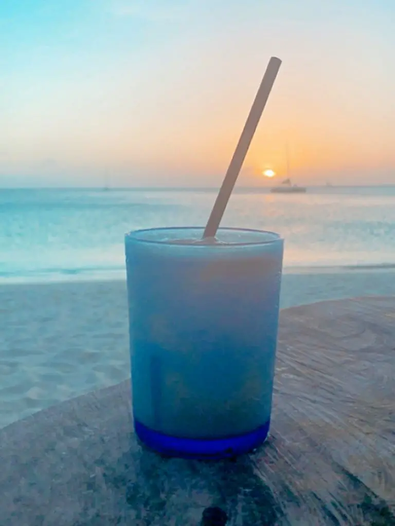 a piña colada in a blue glass sitting on a table on Palm Beach with the ocean and sunset in the background